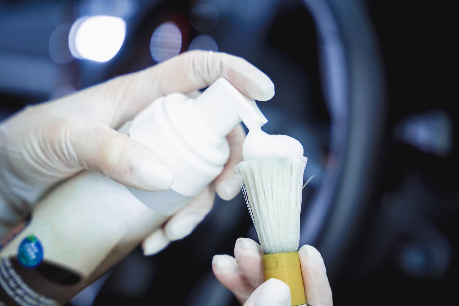 Mãos com luvas aplicam espuma de limpeza numa escova de detalhamento dentro de um carro.